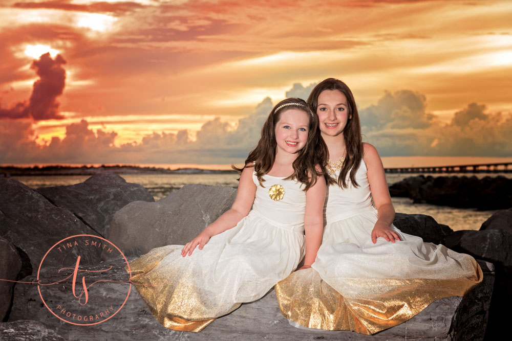 sisters in gold dresses sitting on jetty in destin at sunset