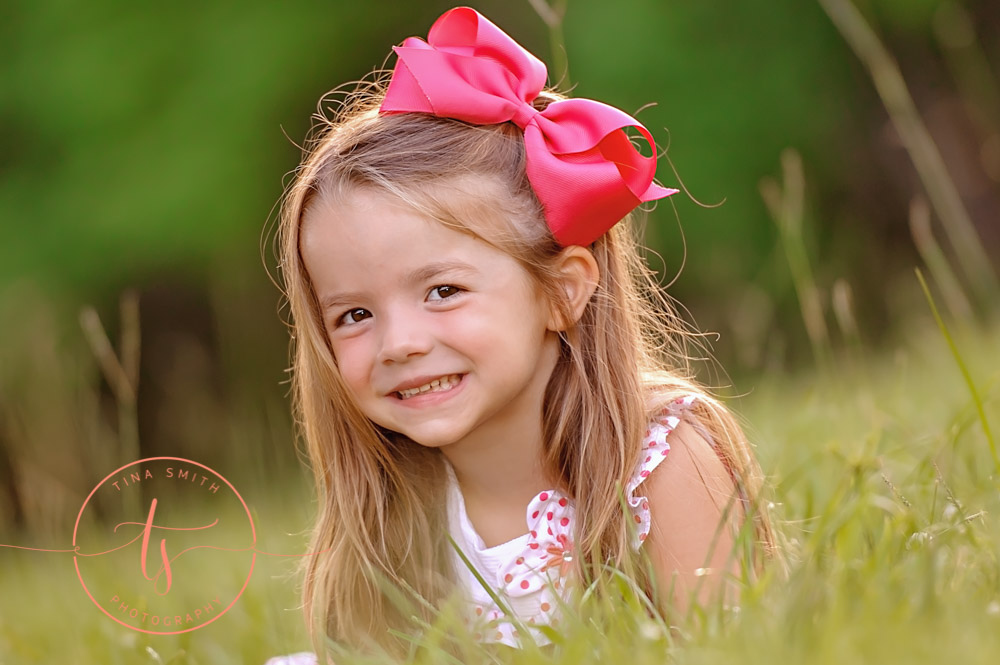 girl in white dress and big pink bow laying in green grass smiling for photographer