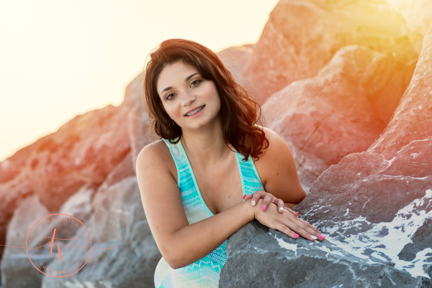 girl in teal shirt posing on rocks on beach in destin for senior portraits at sunset