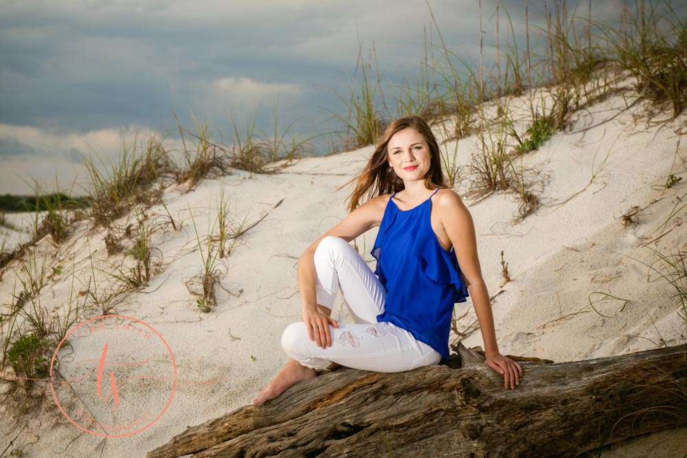 girl posing for destin senior portrait photography in a blue top and white capris sitting on drift wood