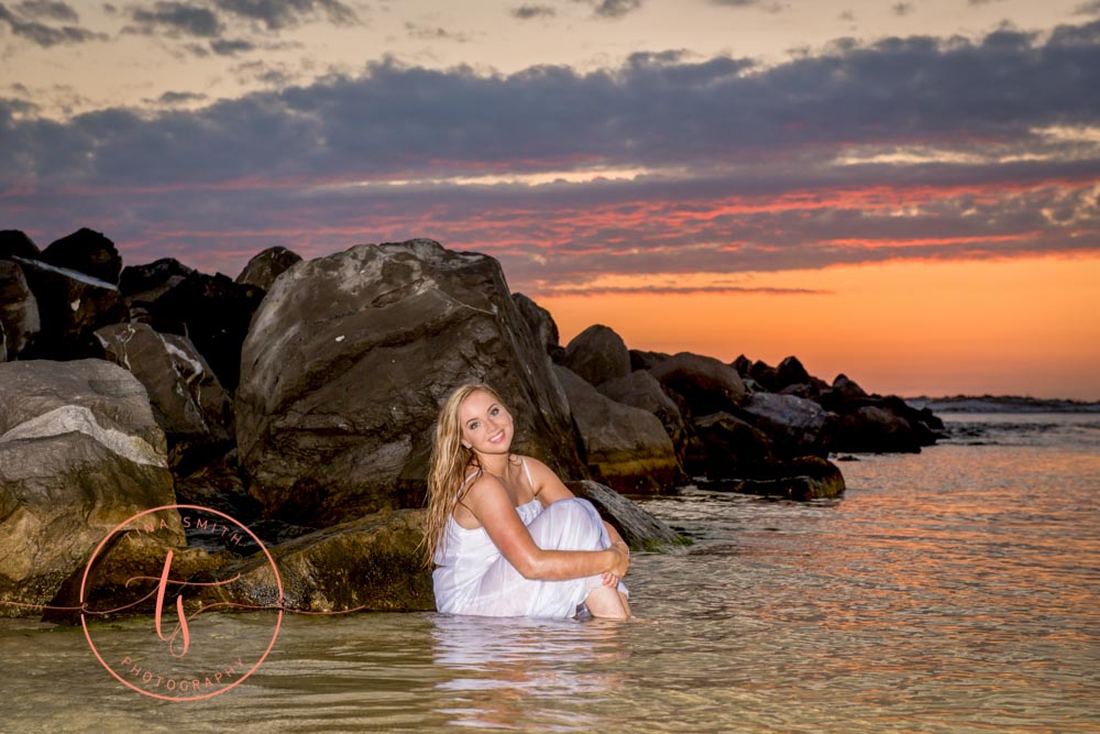 destin senior sitting in water at sunset in white dress posing for destin senior photographer