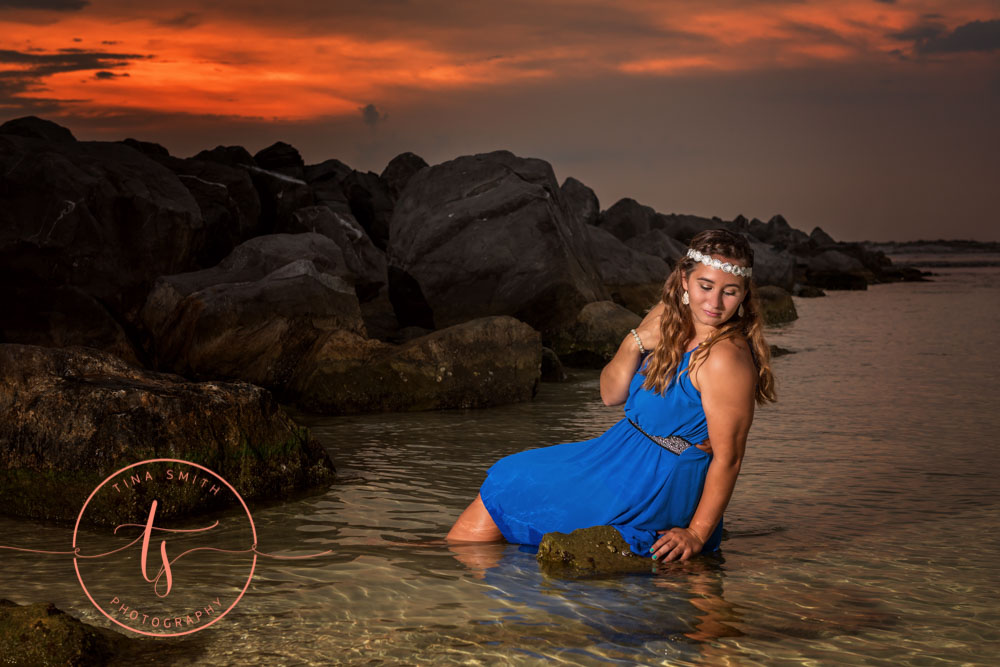 girl posing in water sitting on a rock in formal blue gown for destin senior portraits