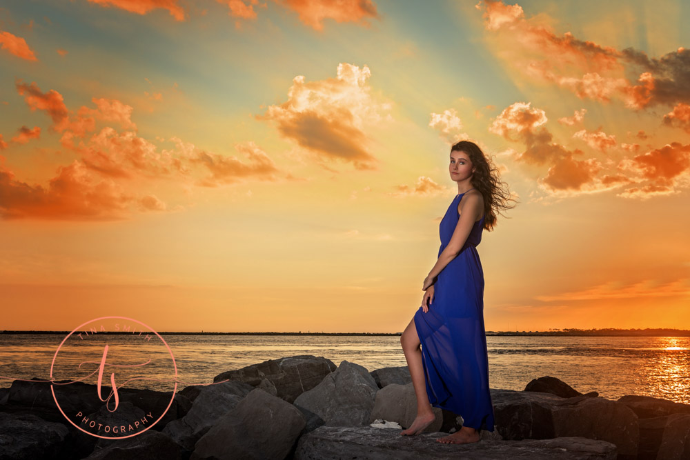 senior girl in blue dress posing on jetties at sunset in destin